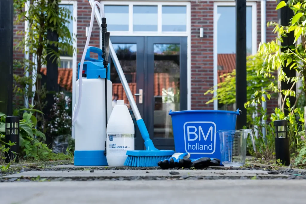 Schoonmaakspullen voor buiten, waaronder een blauwe emmer, een schrobborstel, een witte fles, handschoenen, een maatbeker en een sproeier, liggen op een stenen pad, met op de achtergrond een bakstenen gebouw en groen.