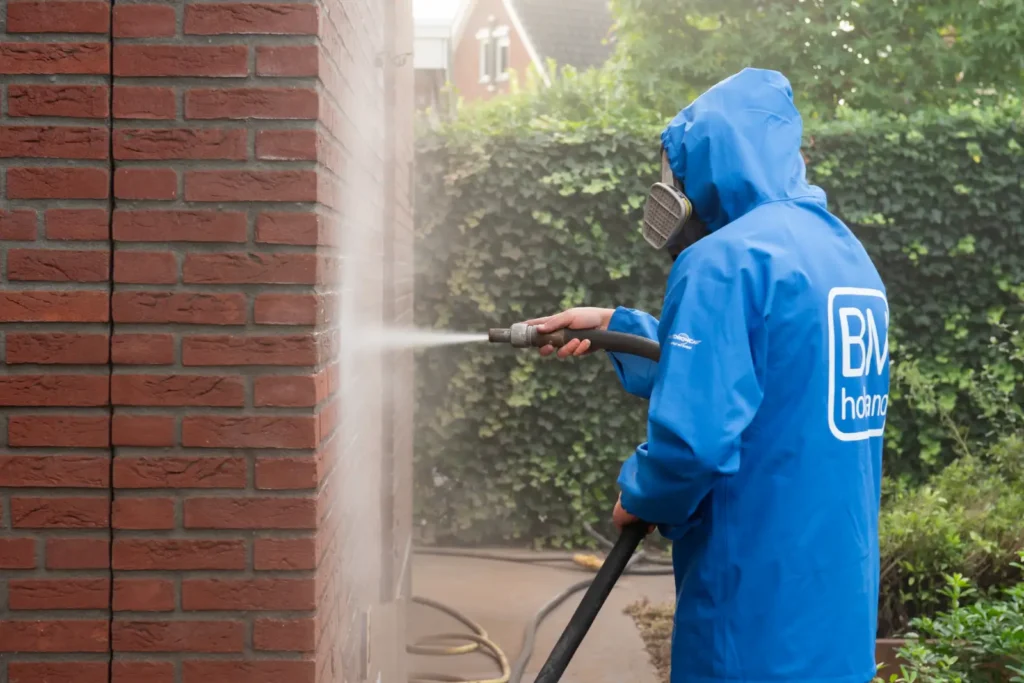 Een persoon in een blauwe jas met capuchon en een gasmasker maakt met een hogedrukreiniger een rode bakstenen muur buiten schoon, terwijl op de achtergrond groen en huizen te zien zijn.
