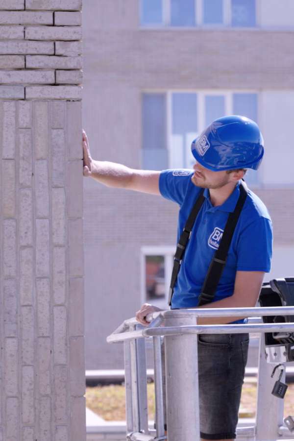 Een bouwvakker met een blauwe bouwhelm en een blauw overhemd inspecteert een bakstenen muur terwijl hij op een hoogwerker buiten een gebouw staat.