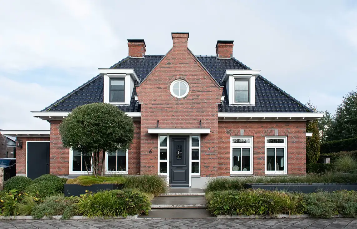 Een bakstenen huis met een donker pannendak, twee schoorstenen, ramen met witte kozijnen, een centraal geplaatste voordeur en een mooi aangelegde voortuin met struiken en bomen.
