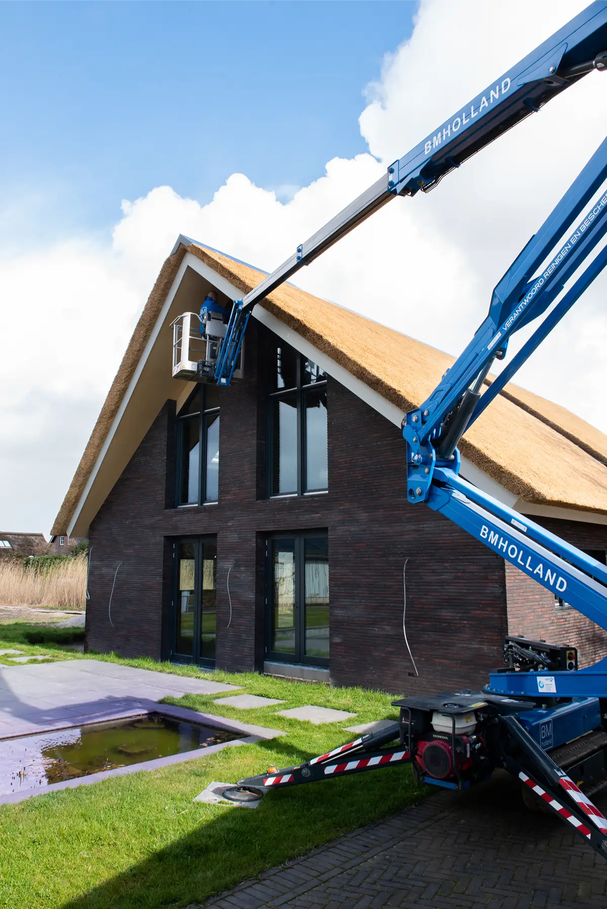 Iemand bedient een blauwe hoogwerker om werkzaamheden uit te voeren op de bovenste verdieping van een moderne woning met een rieten dak en grote ramen.