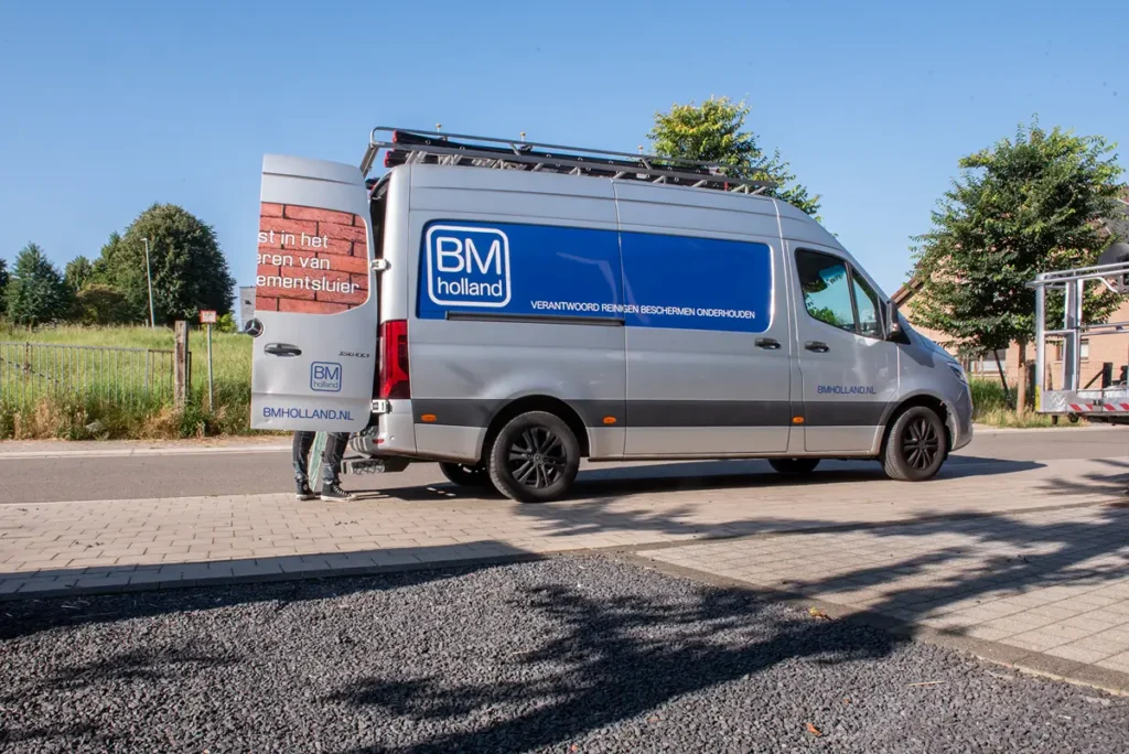 Een zilverkleurig busje van BM Holland met een blauw logo staat op straat geparkeerd. De achterdeur staat open en achterin is gedeeltelijk een persoon te zien. Op de achtergrond staan bomen en is een gebouw te zien.