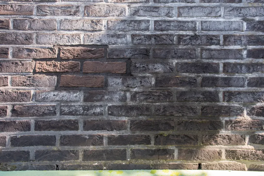 Een bakstenen muur met overwegend donkere stenen, hier en daar wat lichtere stenen bovenaan, en een stukje lichter gekleurde mortel. Het zonlicht werpt gedeeltelijke schaduwen over de muur.