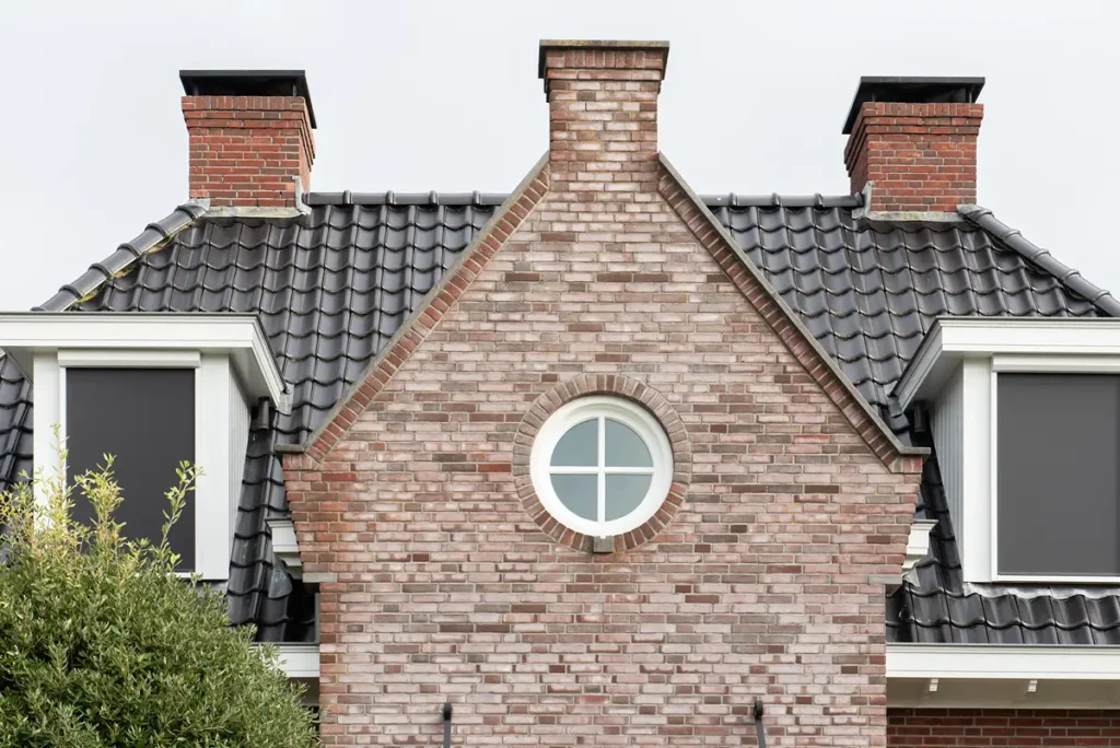 Gevel van een bakstenen huis met twee schoorstenen, een zwart pannendak, twee rechthoekige ramen aan elke kant en een rond raam in het midden. Linksonder zijn gedeeltelijk struiken te zien. De lucht is bewolkt.
