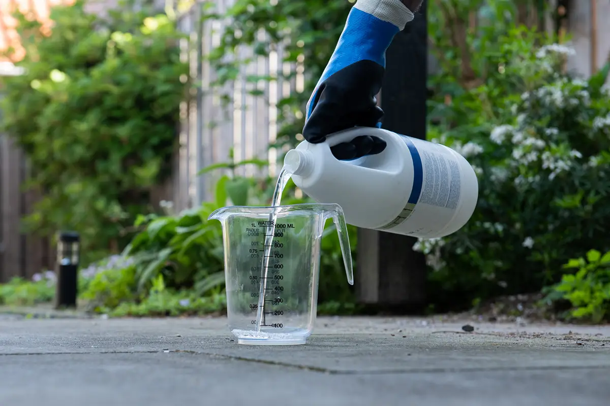 Een persoon met een blauwe handschoen giet buiten vloeistof uit een witte plastic fles in een doorzichtige maatbeker, met groene planten op de achtergrond.