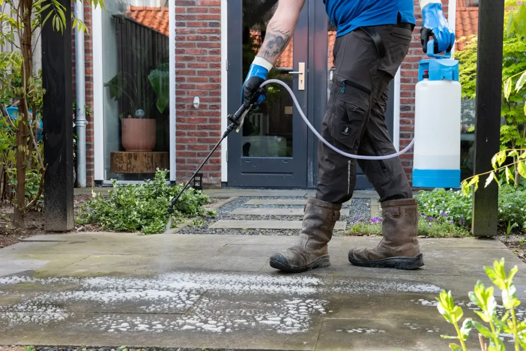 Iemand die handschoenen en laarzen draagt, maakt met een sproeier een terras schoon met zeepschuim, buiten een huis met een glazen deur en bakstenen muren.