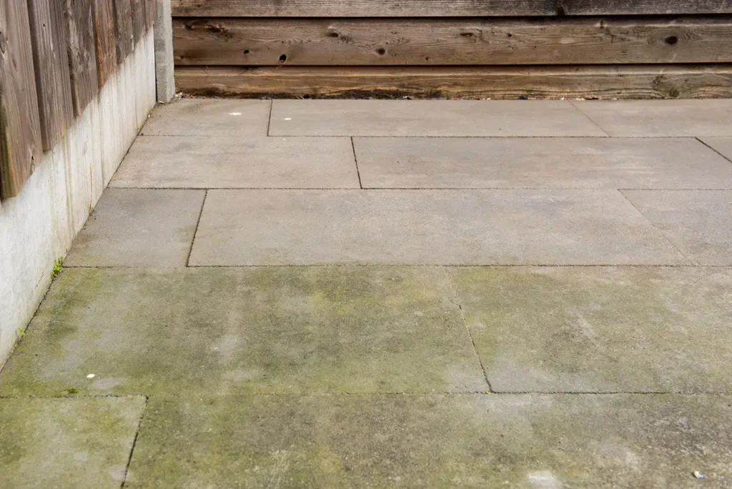 Betonnen terrasplaten met hier en daar wat groen mos of algen aan de onderkant, aan de linkerkant en op de achtergrond omzoomd door een houten schutting, waardoor een hoek van een buitenruimte ontstaat.
