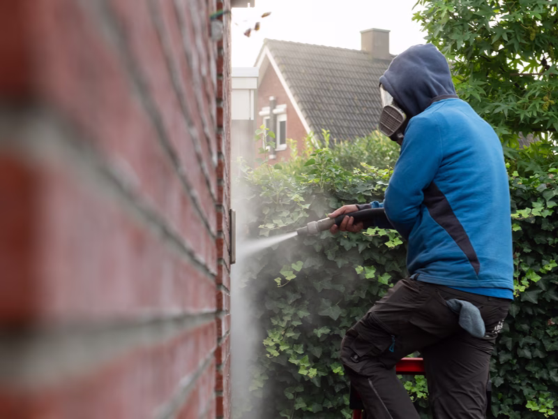 Een persoon in een blauwe hoodie en een mondkapje maakt met een hogedrukreiniger een bakstenen muur buiten schoon; op de achtergrond zijn groene struiken en een huis te zien.