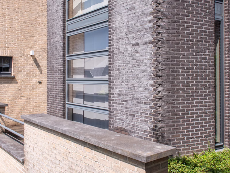 Gevel van een modern gebouw met lichte en donkere bakstenen muren, grote verticale ramen met horizontale kozijnen, en een lage bakstenen muur op de voorgrond, met aan de rechterkant groen zichtbaar.