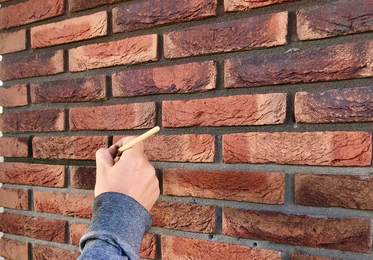 Iemand met een blauwe mouw tekent of trekt met een potlood lijnen op een rode bakstenen muur.