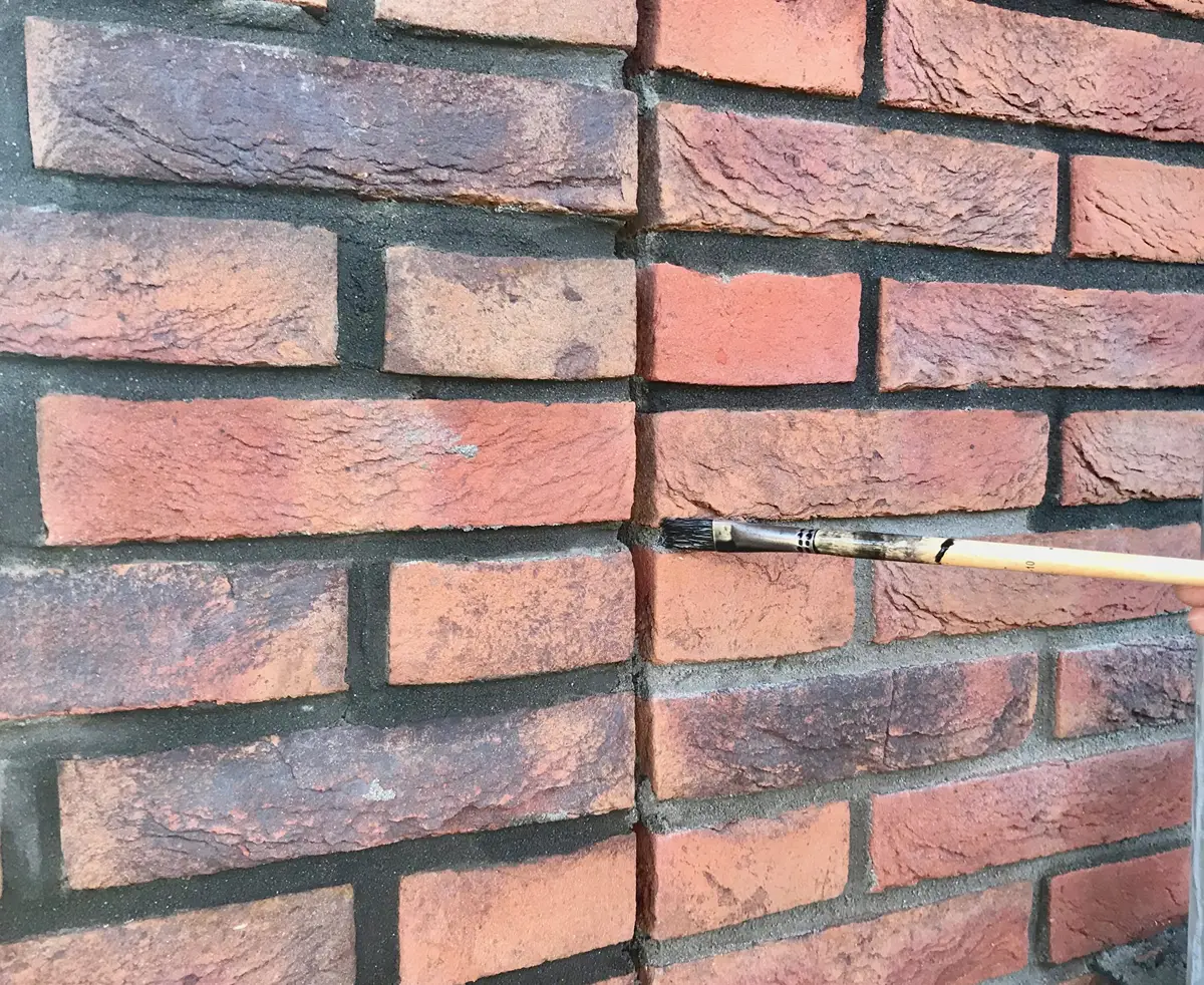 Met een kwast wordt donkere verf aangebracht op de voegen tussen de roodbruine bakstenen van een muur, waardoor de voegen worden geaccentueerd en het geheel een uniforme uitstraling krijgt.
