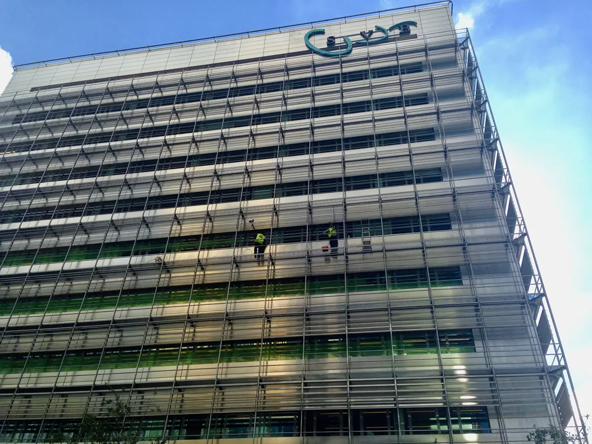 Twee werknemers in veiligheidsuitrusting maken de ramen schoon van een hoog, modern gebouw met horizontale metalen lamellen en groen glas. Het gebouw heeft een logo in de rechterbovenhoek en steekt af tegen een blauwe lucht.