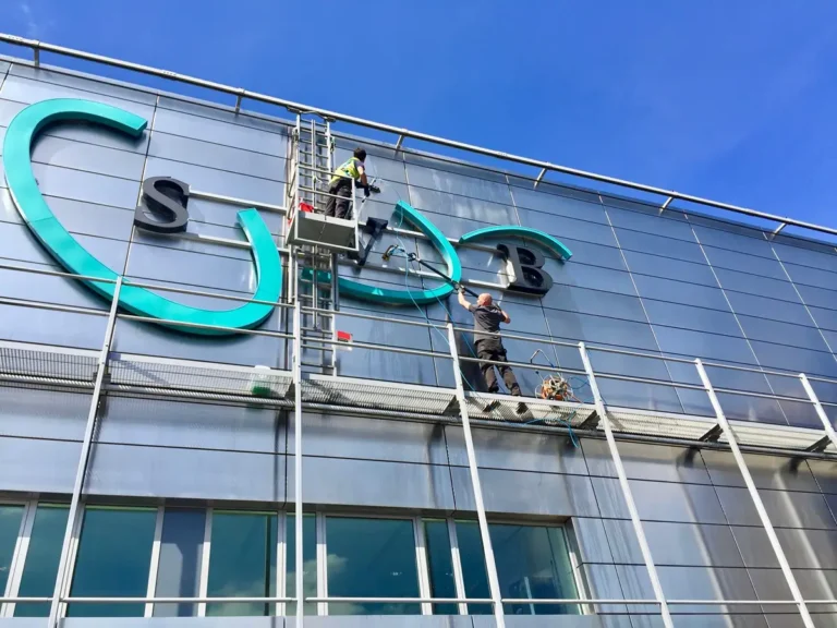 Twee werknemers op hangende platforms zijn bezig met het schoonmaken of onderhouden van de buitenkant van een modern glazen gebouw, vlakbij grote turquoise en zwarte borden, onder een strakblauwe hemel.
