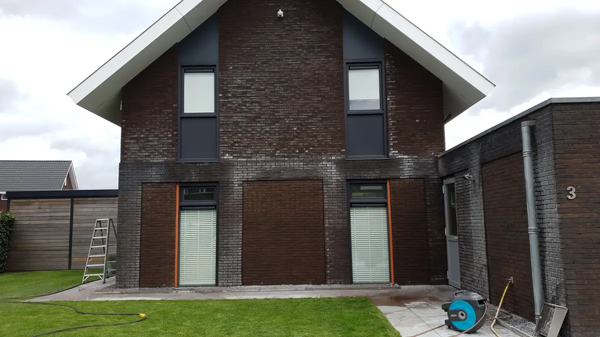 Een bakstenen huis met twee verdiepingen, grote ramen en gesloten jaloezieën. Aan de linkerkant leunt een zilveren ladder, en aan de rechterkant staat een blauwe slanghaspel. De lucht is bewolkt en het huis staat op een grasveld.