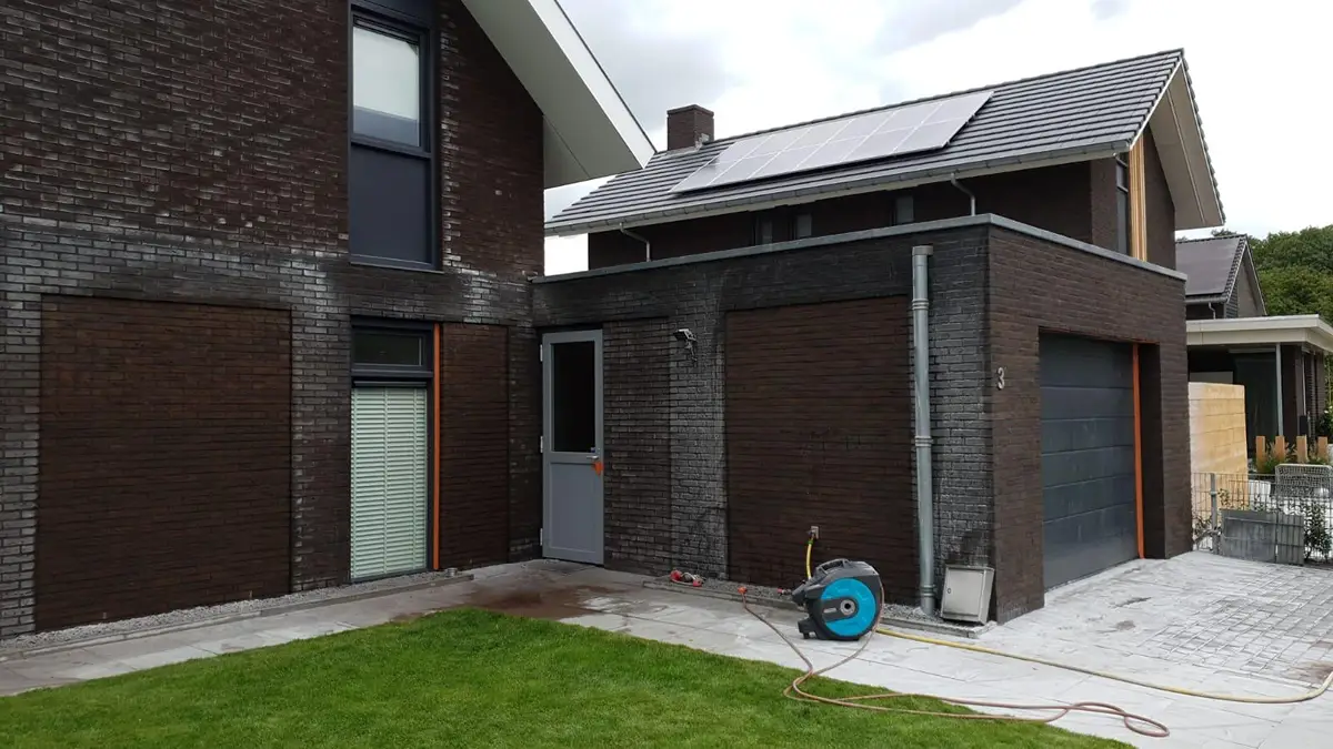 Een moderne bakstenen woning met grote ramen, zonnepanelen op het dak en een garage. Op de oprit en de muren zijn een slanghaspel en natte plekken te zien, wat erop wijst dat er onlangs is schoongemaakt.