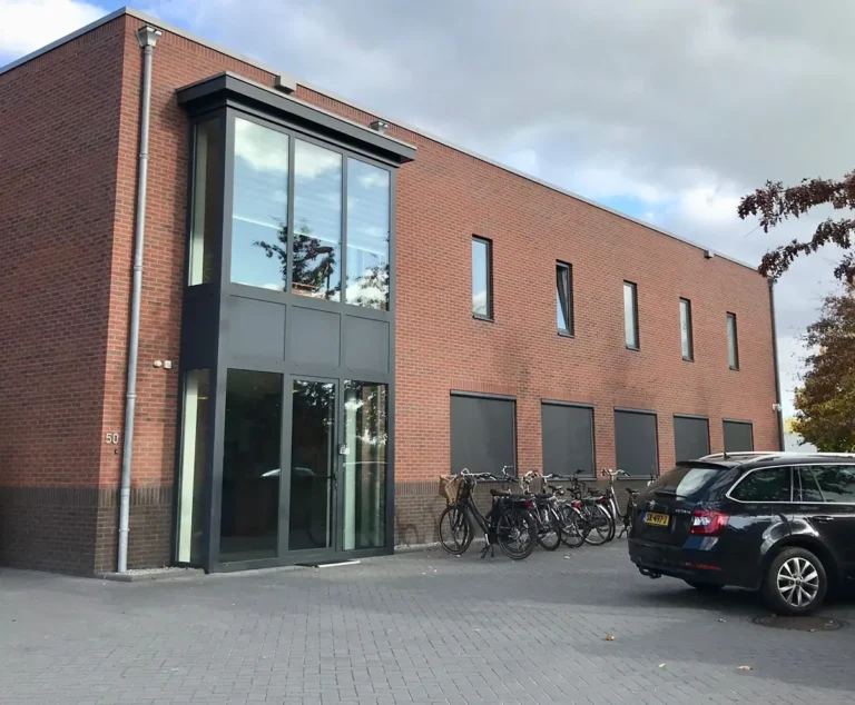 Een modern bakstenen gebouw met grote ramen en een glazen entree. Tegen de muur staan verschillende fietsen geparkeerd, en vlakbij staat een zwarte auto geparkeerd op een verharde parkeerplaats onder een bewolkte hemel.