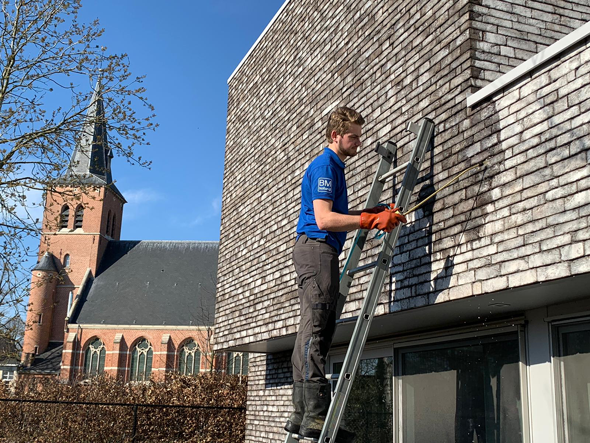 Een man in werkkleding en handschoenen staat op een ladder en maakt de buitenmuur van een modern gebouw schoon. Op de achtergrond is onder een helderblauwe hemel een kerk met een hoge toren te zien.