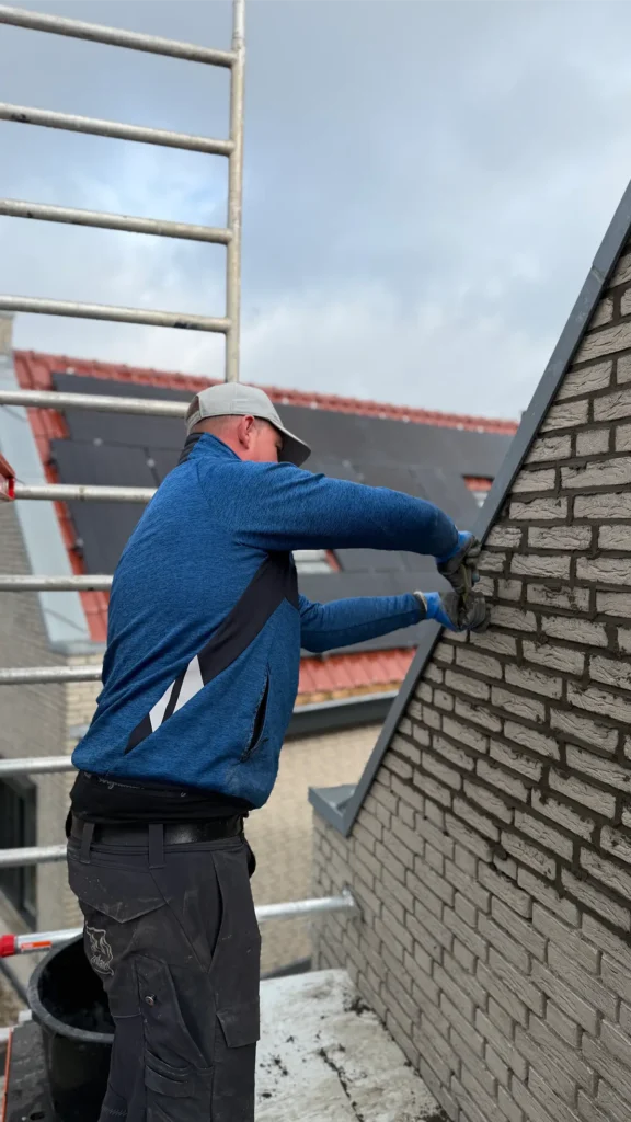 Een persoon in een blauw jasje en een pet is bezig met een bakstenen muur naast een ladder; het lijkt erop dat hij metsel- of reparatiewerkzaamheden uitvoert aan de zijkant van een gebouw, met op de achtergrond daken bedekt met dakpannen.