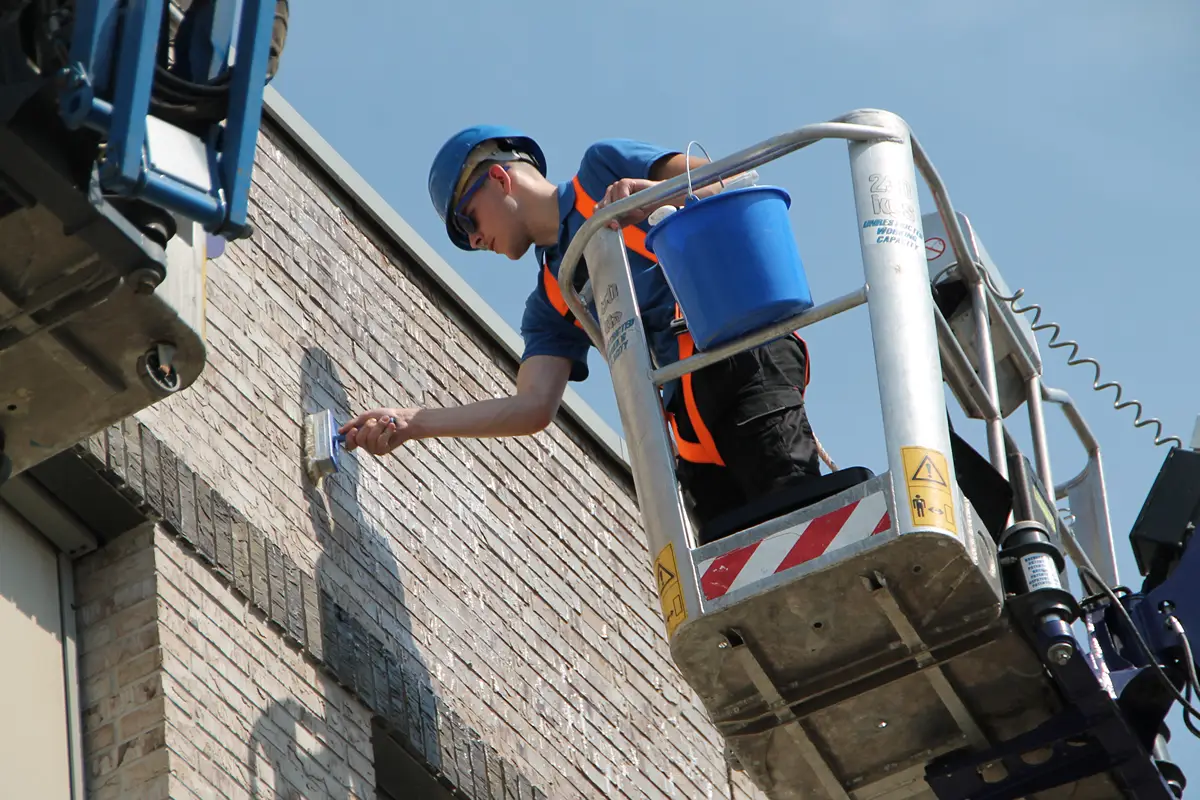 Een werknemer met een blauwe veiligheidshelm en een veiligheidsharnas schildert een bakstenen muur terwijl hij in een hoogwerker staat. Naast hem hangt een blauwe emmer aan de reling, en op de achtergrond is de lucht helder.