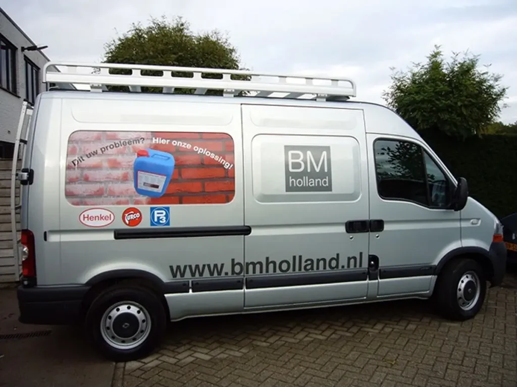 Een wit bestelbusje van BM Holland staat geparkeerd in een straat. Op de zijkant van het busje staan het bedrijfslogo, een website-URL en een advertentie voor schoonmaakproducten. Op de achtergrond zijn bomen en gebouwen te zien.