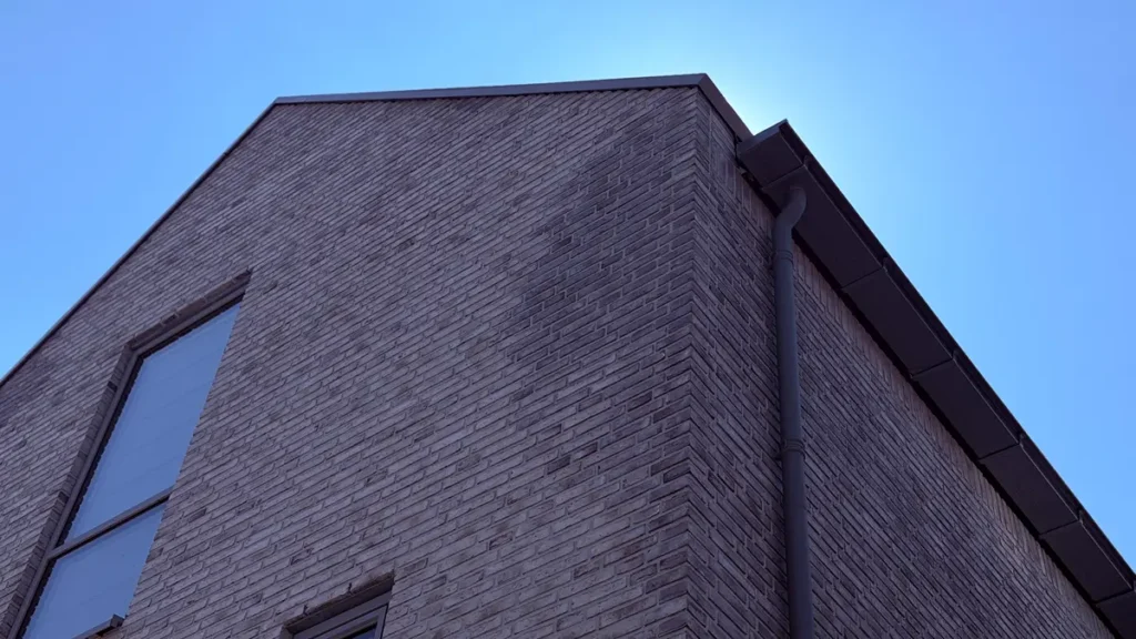 Een close-up van de hoek van een bakstenen gebouw tegen een helderblauwe lucht, met een raam en een regengoot.