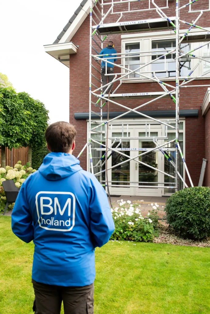 Een man in een blauw BM Holland-jack staat op een gazon en kijkt naar een andere werker die op een steiger staat voor een bakstenen huis met witte raamkozijnen.
