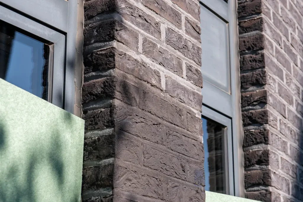 Close-up van de hoek van een donker bakstenen gebouw met twee ramen, grijze sierlijsten en groen bouwmateriaal dat delen van de onderste muren bedekt. Het zonlicht werpt schaduwen op het bakstenen oppervlak.