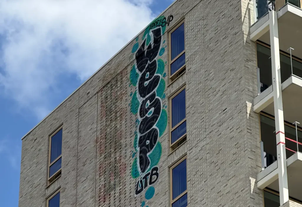 Op de bovenste muur van een beige bakstenen gebouw, vlakbij verschillende ramen en balkons, is verticaal graffiti aangebracht met het woord JESSO in blauwgroen en zwart. Er zijn een blauwe lucht en wolken te zien.