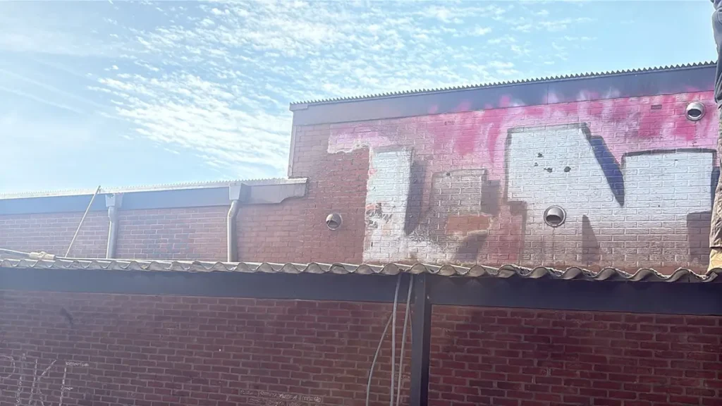 Graffiti in vervaagde roze en witte verf bedekt een deel van de muur van een bakstenen gebouw onder een gedeeltelijk bewolkte hemel, met een golfplaten dak op de voorgrond.