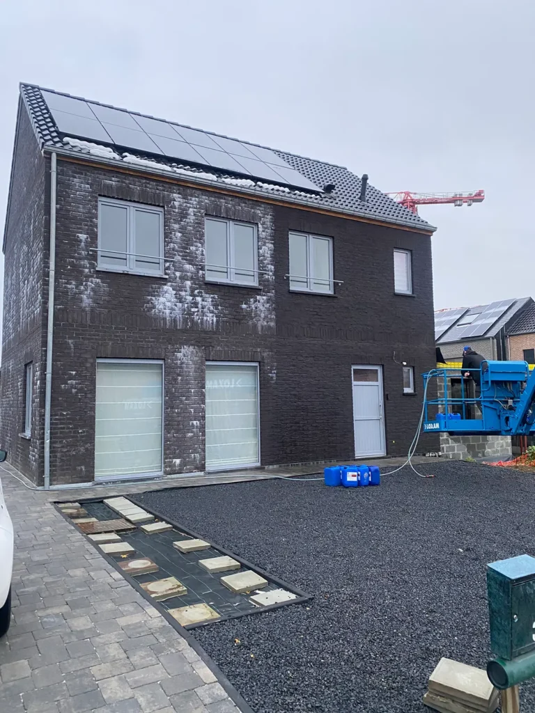 Een moderne woning met twee verdiepingen, opgetrokken uit donkere baksteen, met zonnepanelen op het dak, witte vlekken op de muren en bouwmachines die ervoor geparkeerd staan. De tuin is nog niet helemaal aangelegd en bestaat uit grind en straatstenen.