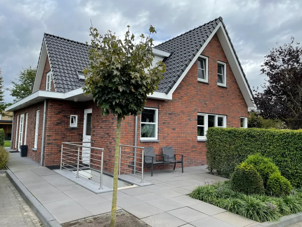 Een moderne bakstenen woning met een zadeldak, witte sierlijsten, een kleine veranda met een metalen leuning, twee stoelen en een boom ervoor, omringd door keurig gesnoeide struiken en met boven zich een bewolkte hemel.