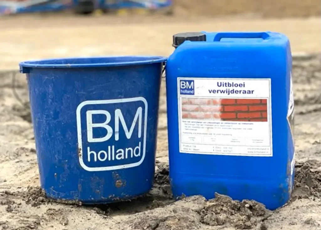 Een blauwe emmer van BM Holland en een blauwe container met het opschrift ‘Uitbloei verwijderaar’ staan op zandgrond op een bouwplaats, met op de achtergrond een wazig gebouw en machines.